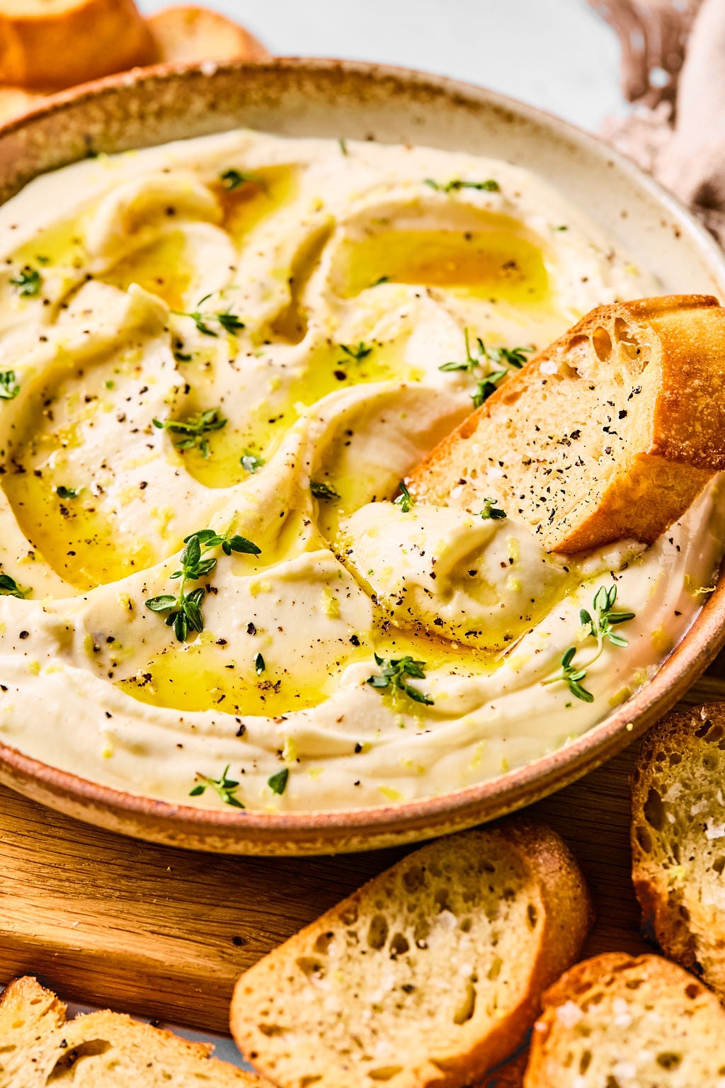 A bowl of whipped ricotta garnished with herbs and black pepper, with a slice of toasted bread dipped in.
