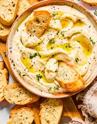 A bowl of whipped ricotta dip garnished with oil, herbs, and black pepper, surrounded by slices of toasted baguette on a wooden board.