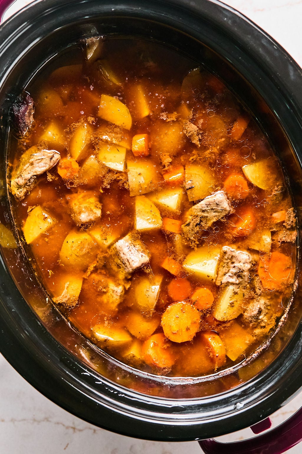A slow cooker filled with beef stew containing chunks of beef, potatoes, carrots, and broth.