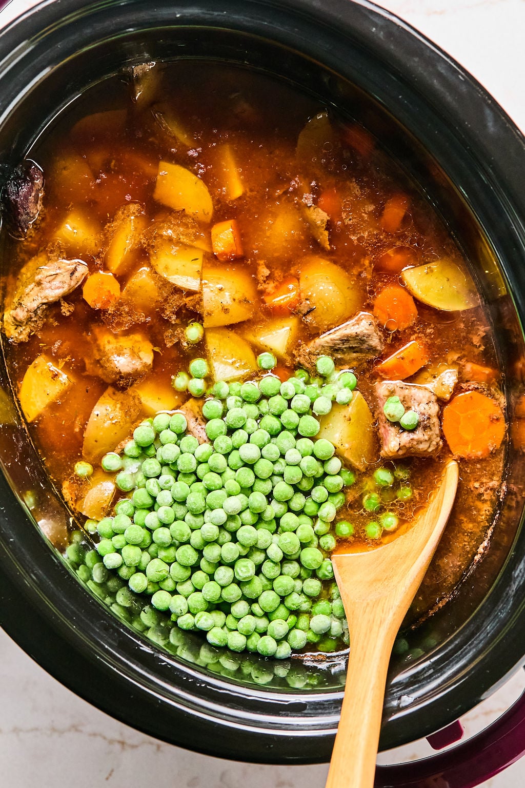 A slow cooker filled with beef stew containing chunks of beef, potatoes, carrots, broth, and a pile of frozen peas on top, with a wooden spoon resting inside.