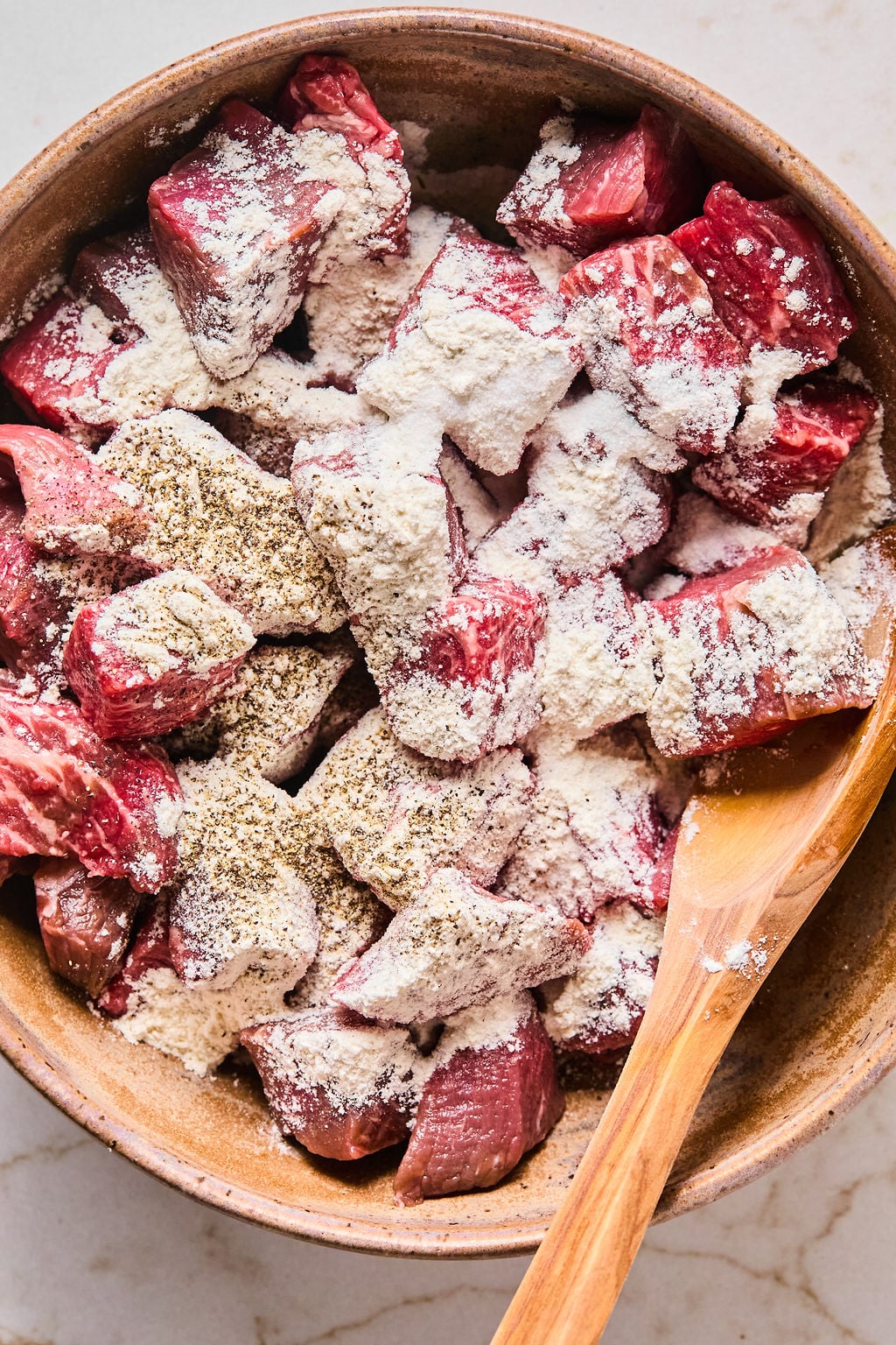 Chunks of raw beef in a brown bowl coated with flour and black pepper, with a wooden spoon resting in the bowl.