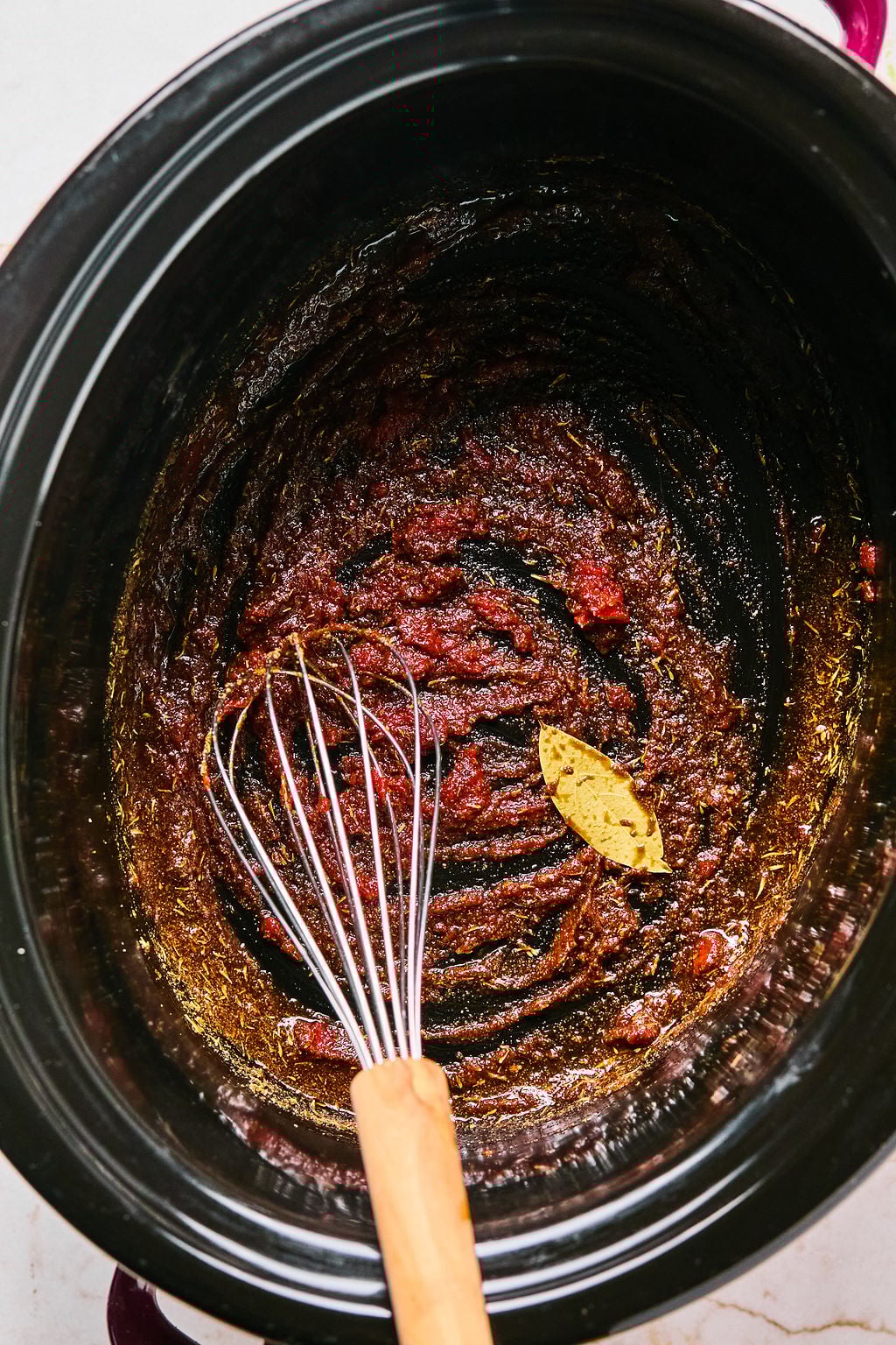 A whisk rests in a slow cooker containing tomato paste and a bay leaf, partially mixed together.