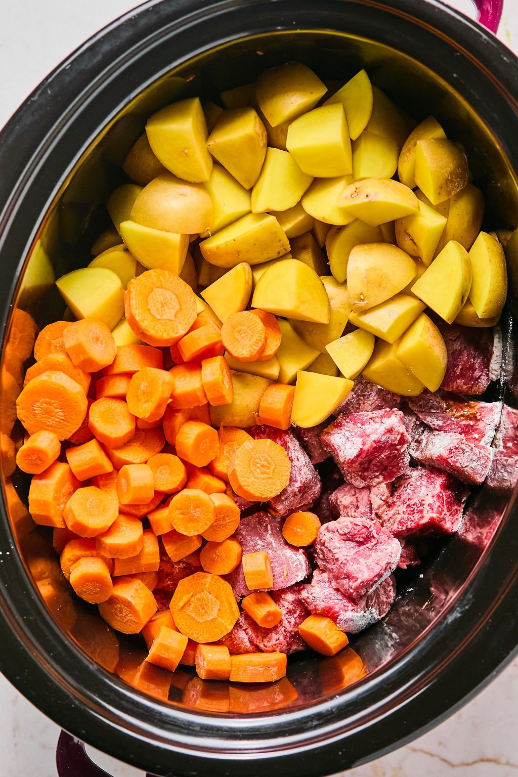 Chopped potatoes, carrots, and raw beef chunks placed in a slow cooker.