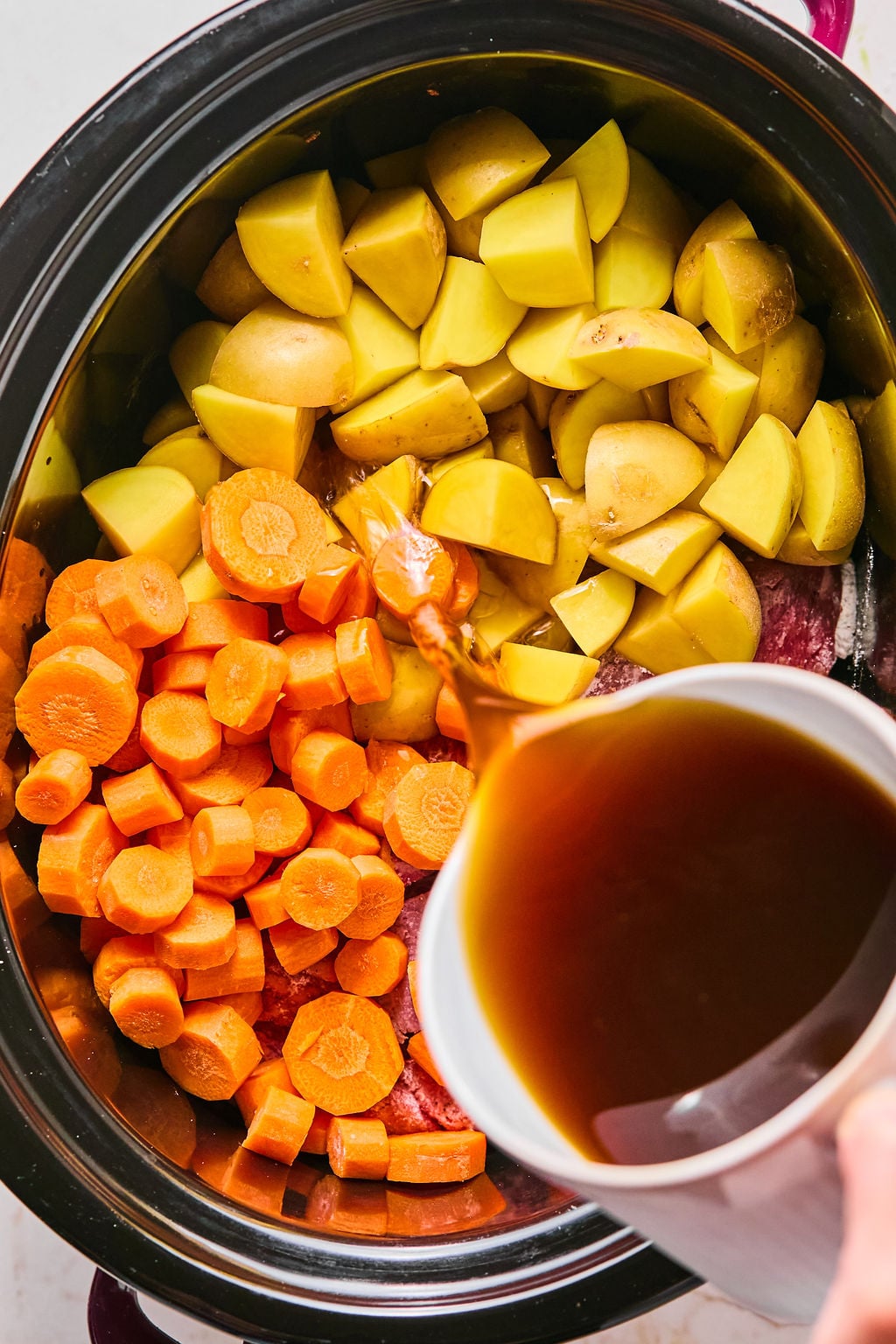 Chopped potatoes and carrots in a slow cooker with broth being poured in from a measuring cup.