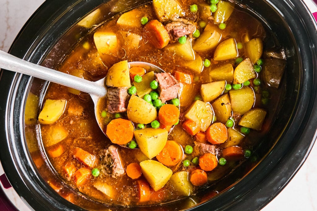 A ladle scoops beef stew with potatoes, carrots, and peas from a slow cooker filled with broth and vegetables.