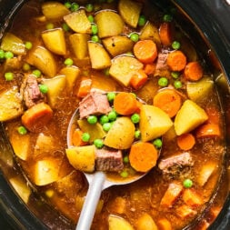 A slow cooker filled chunks of beef, potatoes, carrots, and green peas, with a ladle scooping out a serving.