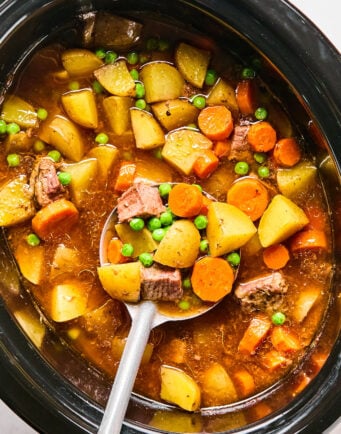 A slow cooker filled chunks of beef, potatoes, carrots, and green peas, with a ladle scooping out a serving.