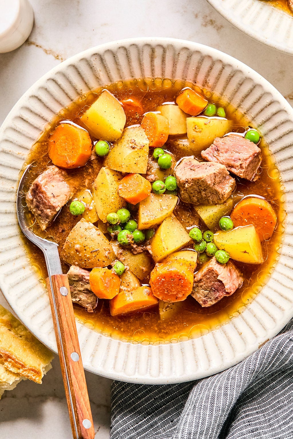 A bowl of beef stew with chunks of beef, potatoes, carrots, and peas in broth, served with a spoon on a white plate.