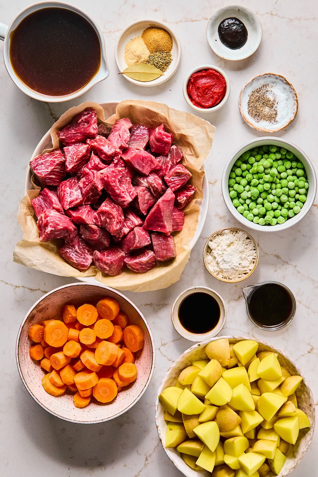 Assorted beef stew ingredients on a countertop, including chunks of raw beef, carrots, potatoes, peas, broth, flour, seasonings, tomato paste, and sauce in bowls and cups.
