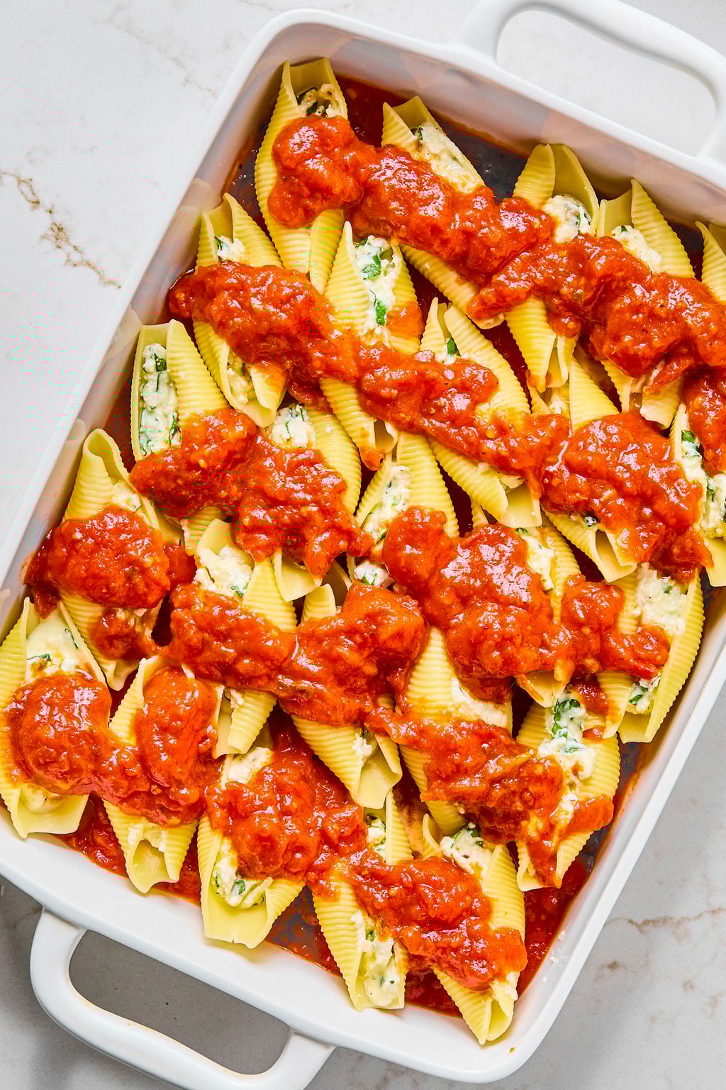 A white baking dish filled with uncooked pasta shells stuffed with ricotta cheese mixture and topped with red tomato sauce.