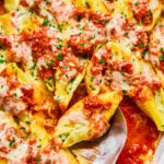 Baked stuffed pasta shells topped with tomato sauce and melted cheese in a white dish, with a metal serving spoon.