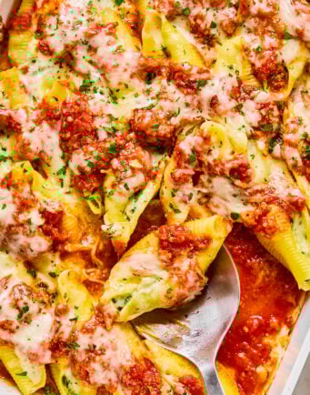 Baked stuffed pasta shells topped with tomato sauce and melted cheese in a white dish, with a metal serving spoon.