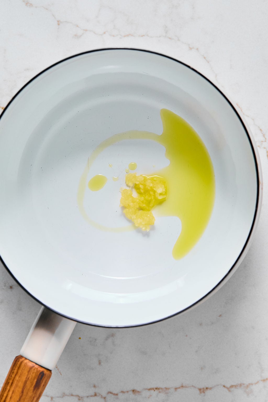 A white pan contains a small amount of olive oil and minced garlic on a white marble surface.