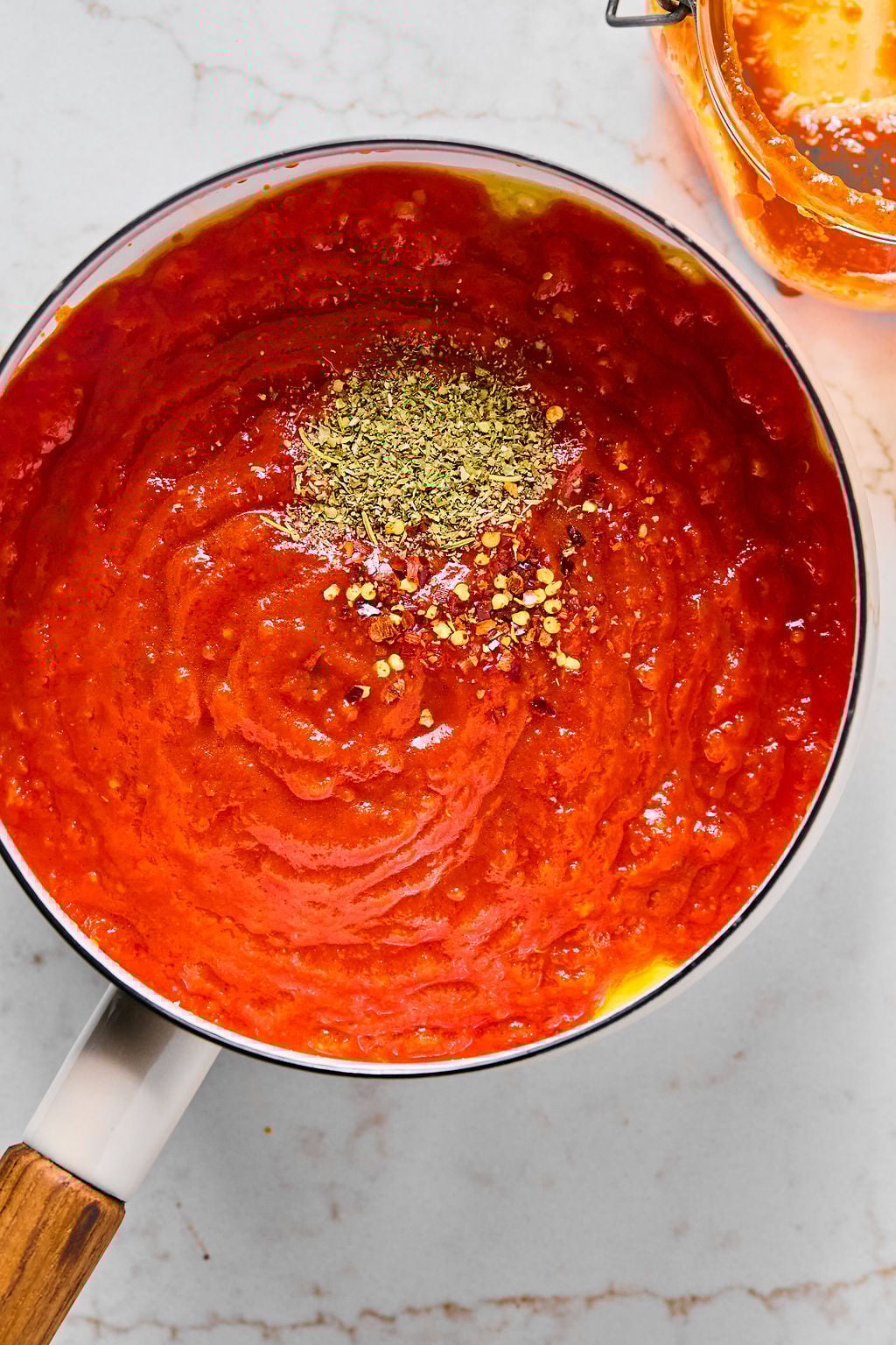 A saucepan filled with tomato sauce, topped with dried oregano and red pepper flakes, sits on a marble surface.