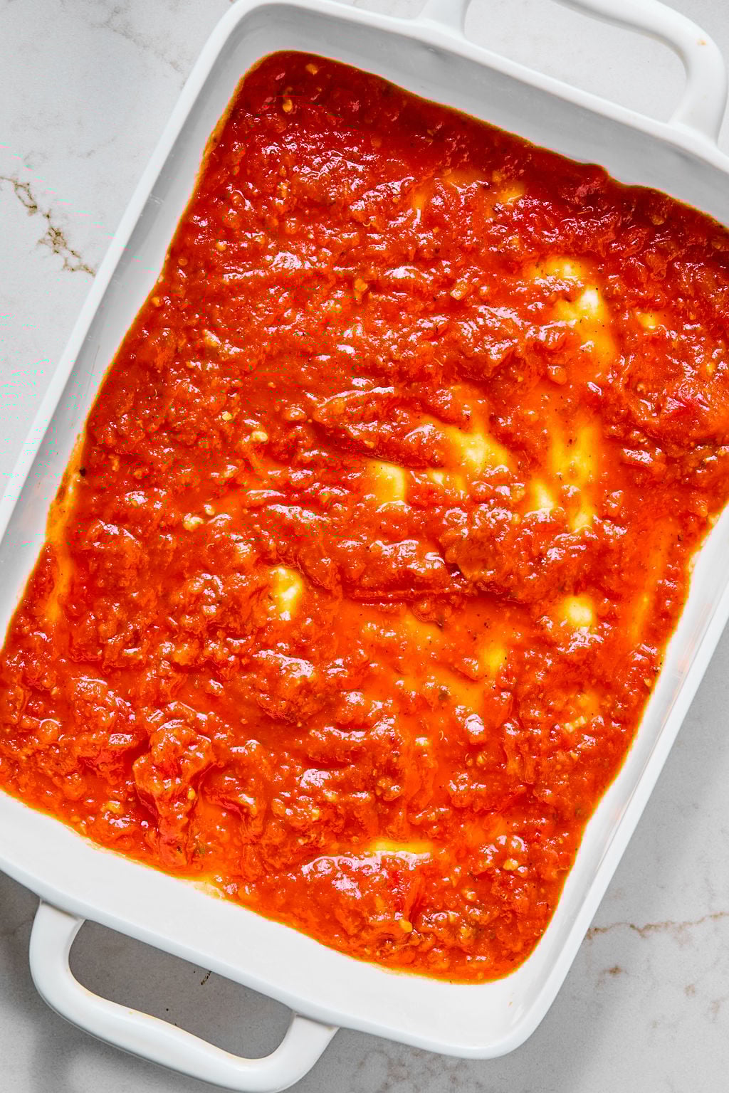 A white rectangular baking dish filled with a layer of red tomato sauce, spread evenly across the bottom.