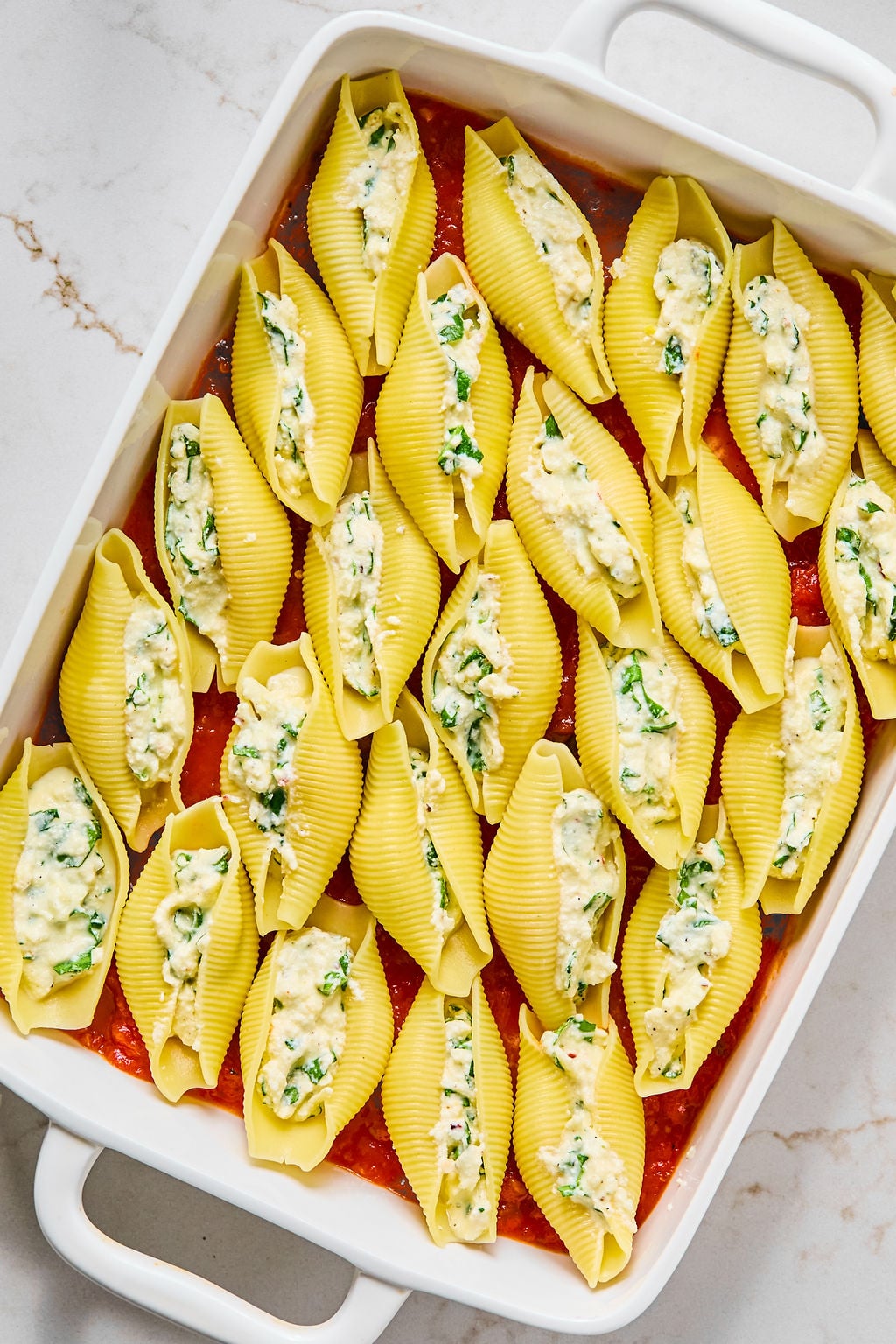 A white baking dish filled with uncooked pasta shells stuffed with a cheese and spinach mixture, arranged over a layer of tomato sauce.