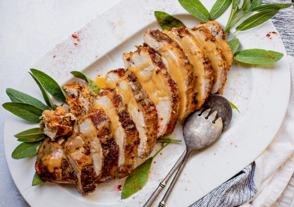 Sliced roasted turkey breast with gravy on a white platter, garnished with fresh sage leaves and served with a large silver fork and spoon.