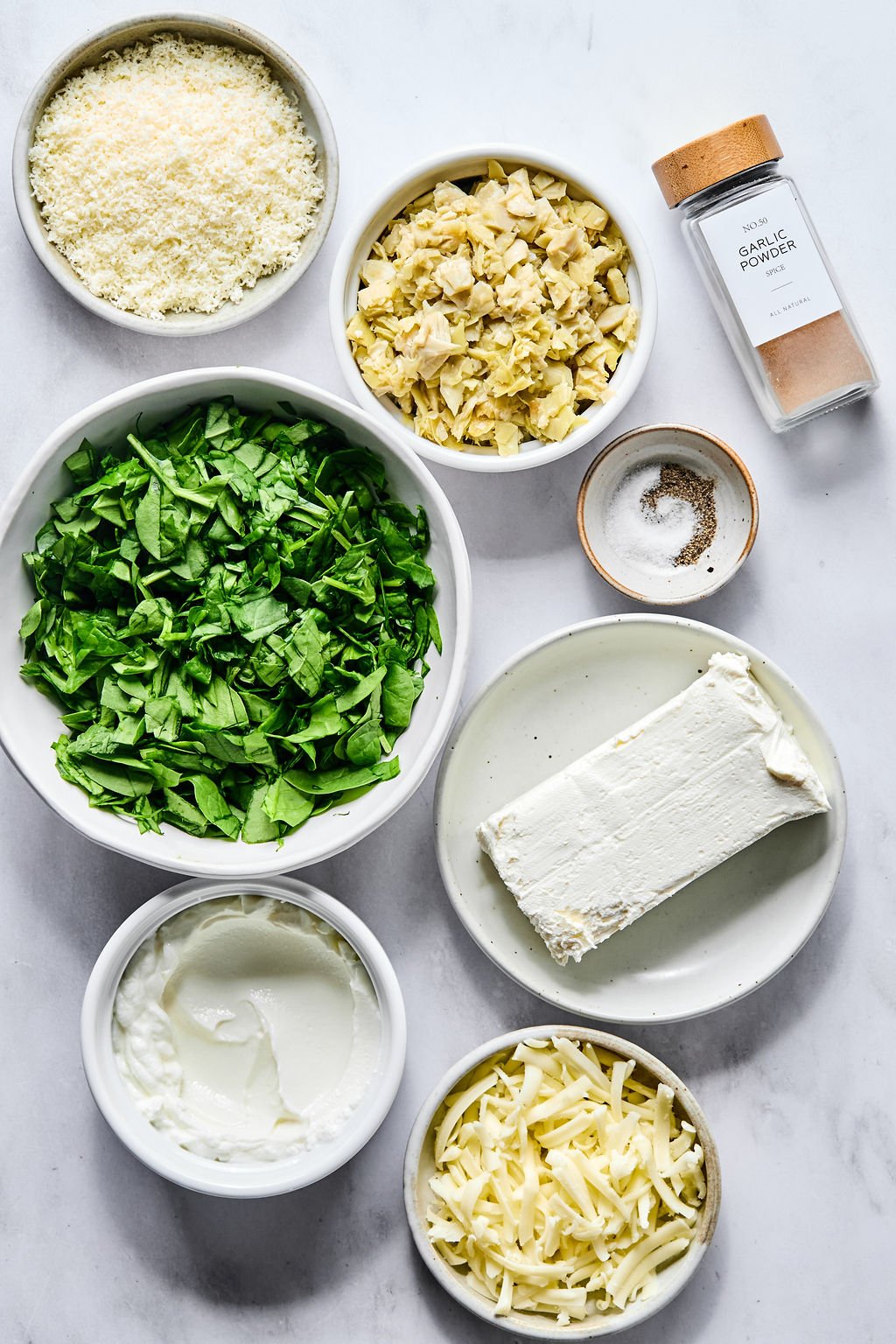 Seven bowls containing grated Parmesan, chopped artichoke hearts, garlic powder, salt and pepper, chopped spinach, cream cheese, shredded mozzarella, and sour cream on a white surface.