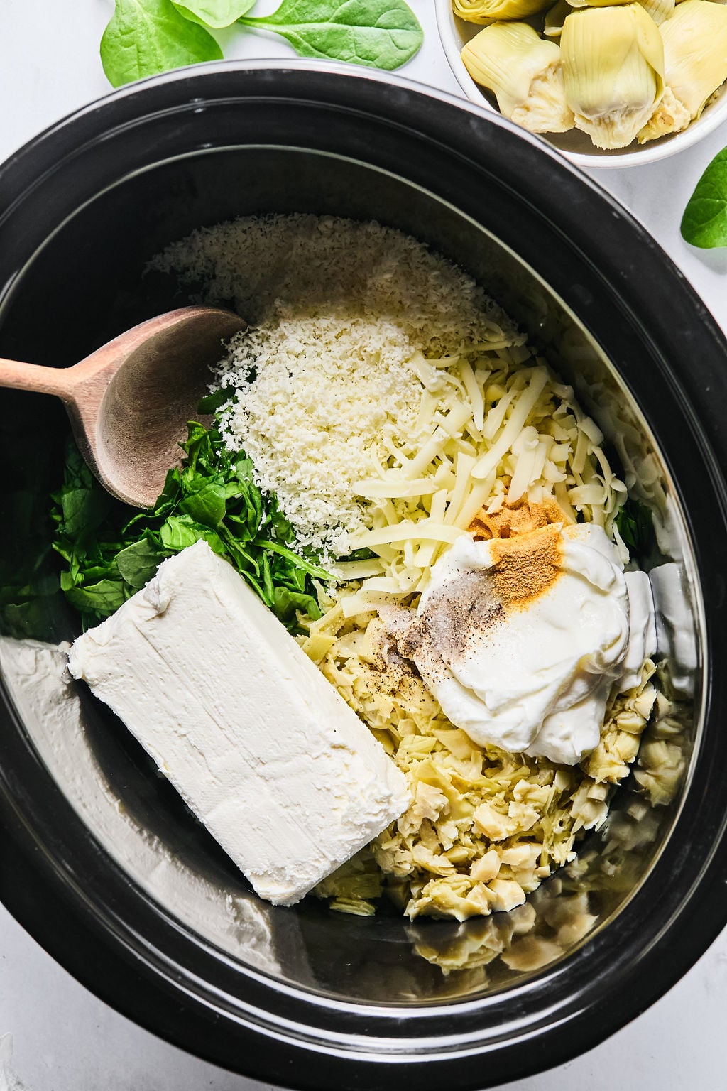 Ingredients for a dip, including cream cheese, shredded cheese, spinach, artichokes, sour cream, and seasonings, are placed in a slow cooker with a wooden spoon.