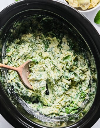 A wooden spoon rests in a slow cooker filled with spinach artichoke dip.