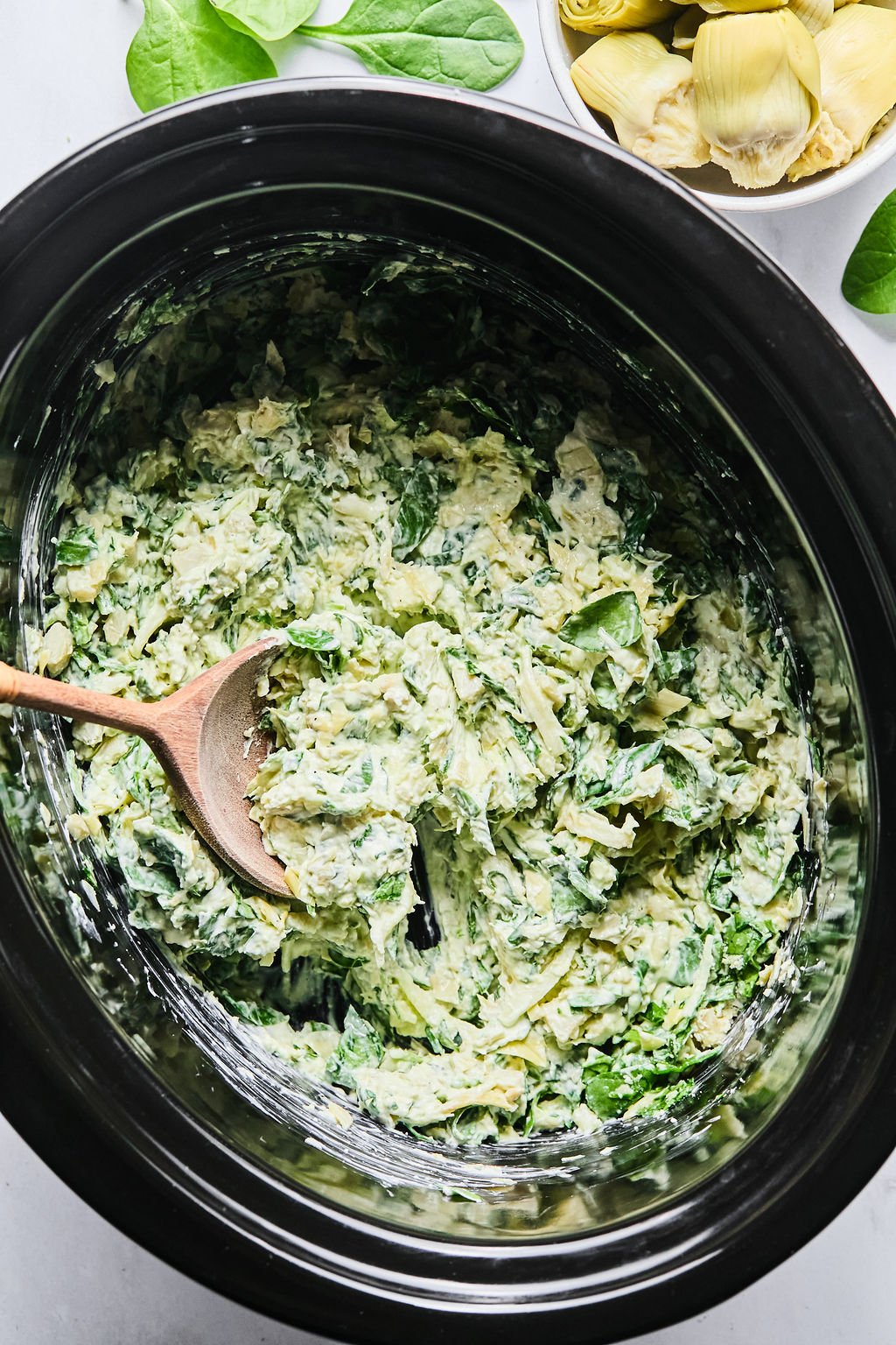 A wooden spoon rests in a slow cooker filled with spinach artichoke dip.