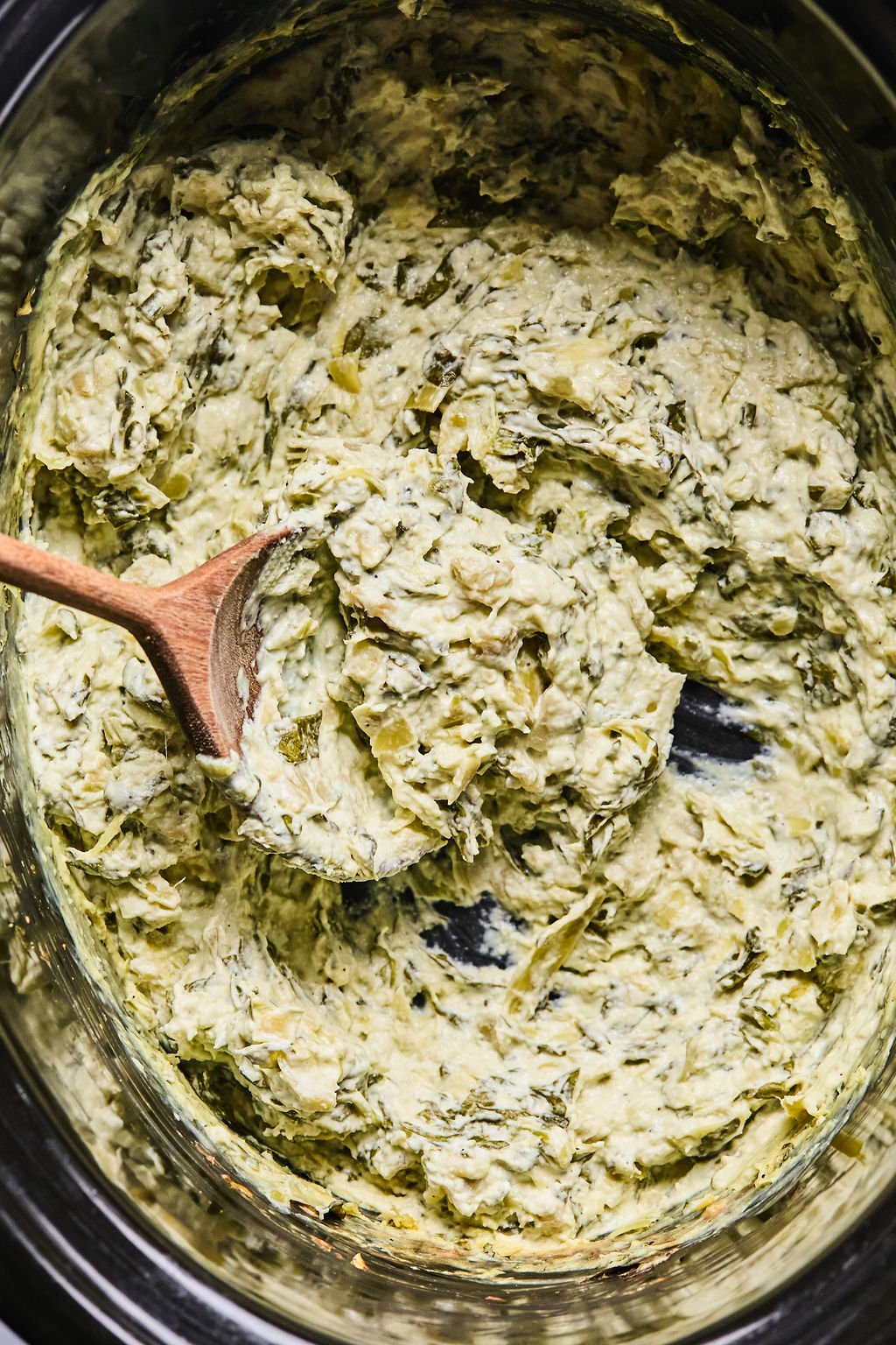 A wooden spoon stirs creamy spinach and artichoke dip inside a slow cooker.