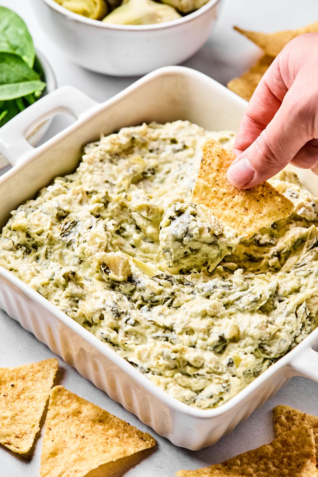 A hand dips a tortilla chip into a baking dish filled with creamy spinach artichoke dip; chips and greens are visible nearby.