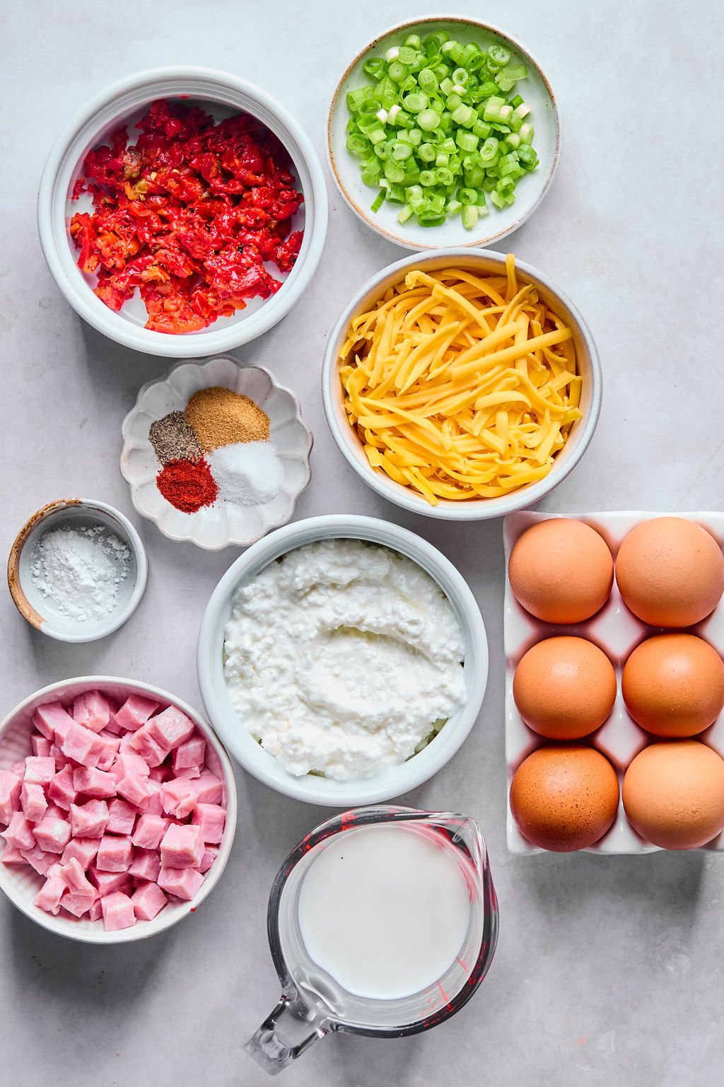 Various ingredients including chopped red peppers, green onions, shredded cheese, eggs, cubed ham, cottage cheese, milk, and assorted spices arranged in bowls on a white surface.