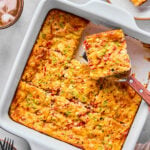 A baked egg casserole with a golden top, cut into squares, with a spatula lifting one piece; plates and drinks are nearby on the table.