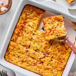A baked egg casserole with a golden top, cut into squares, with a spatula lifting one piece; plates and drinks are nearby on the table.