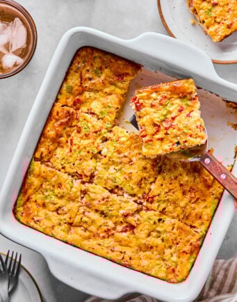 A baked egg casserole with a golden top, cut into squares, with a spatula lifting one piece; plates and drinks are nearby on the table.