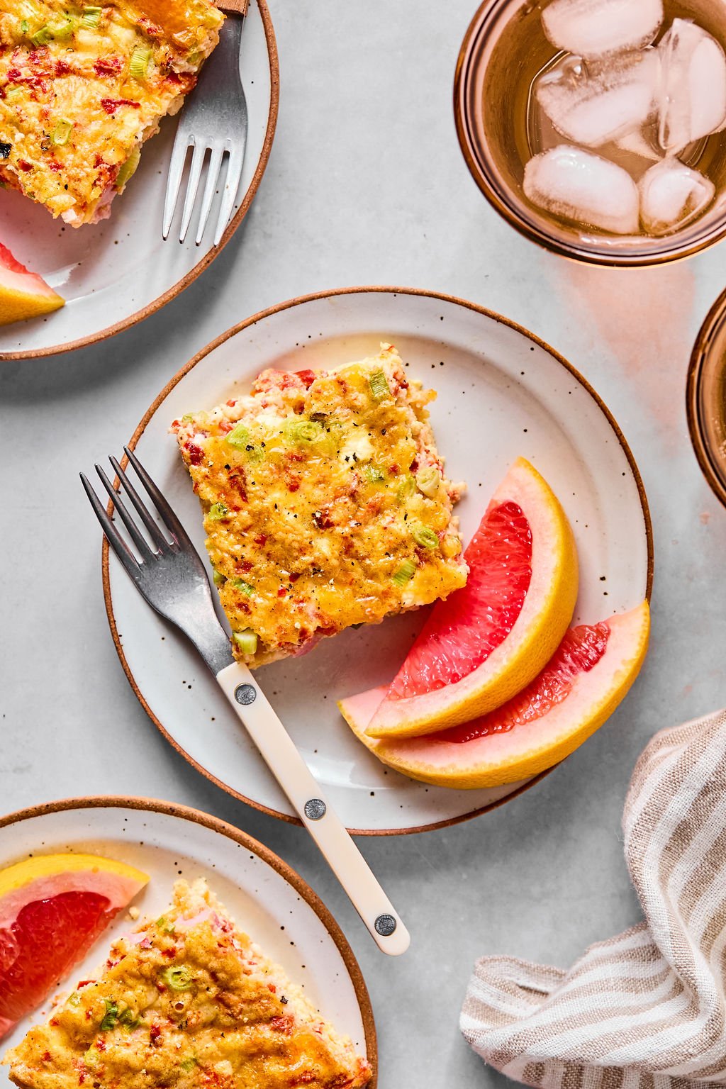 A plate with a slice of baked egg casserole, two grapefruit wedges, a fork, and a glass of iced drink on a light surface.