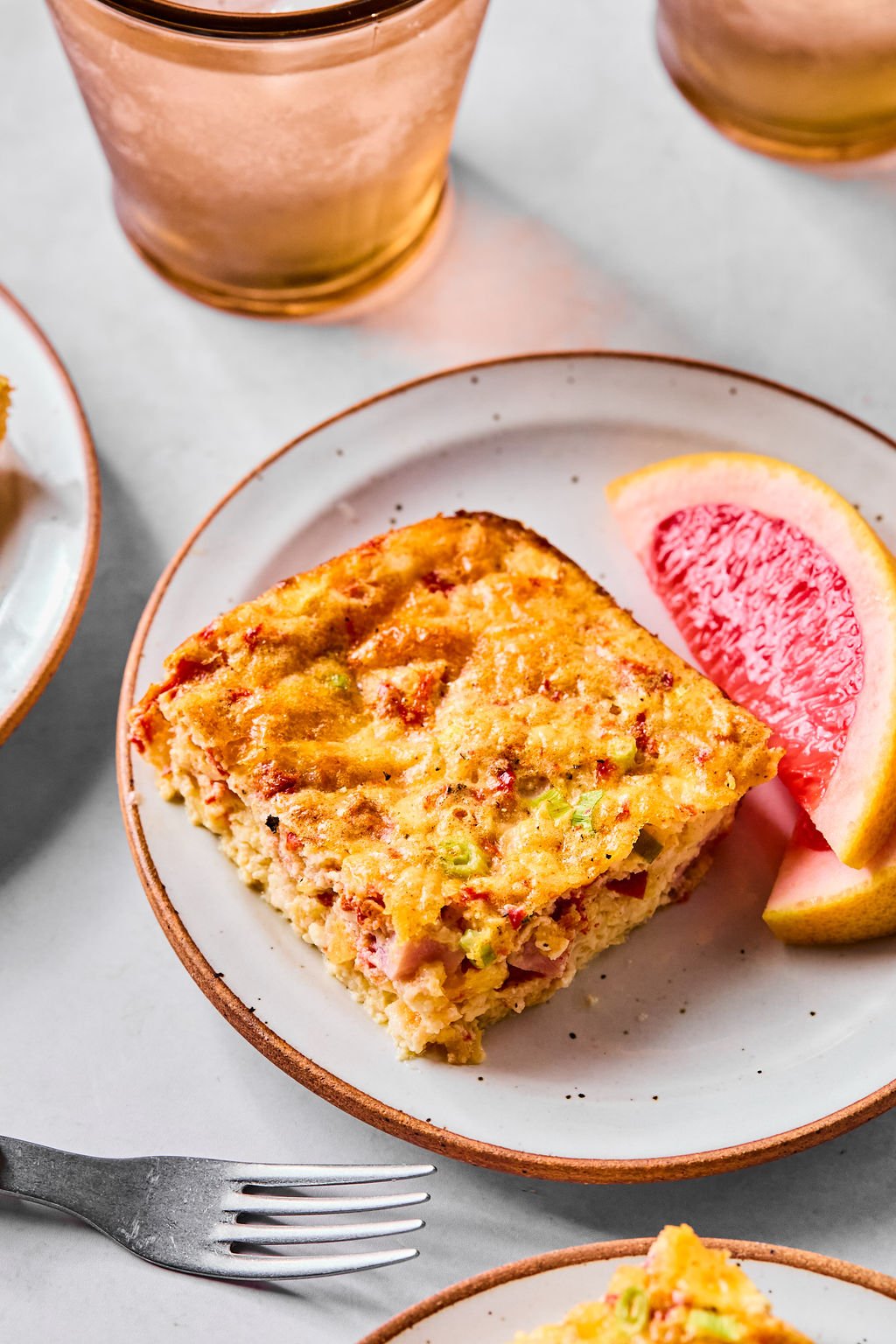 A slice of baked egg casserole with visible vegetables and meat, served on a plate with grapefruit wedges.