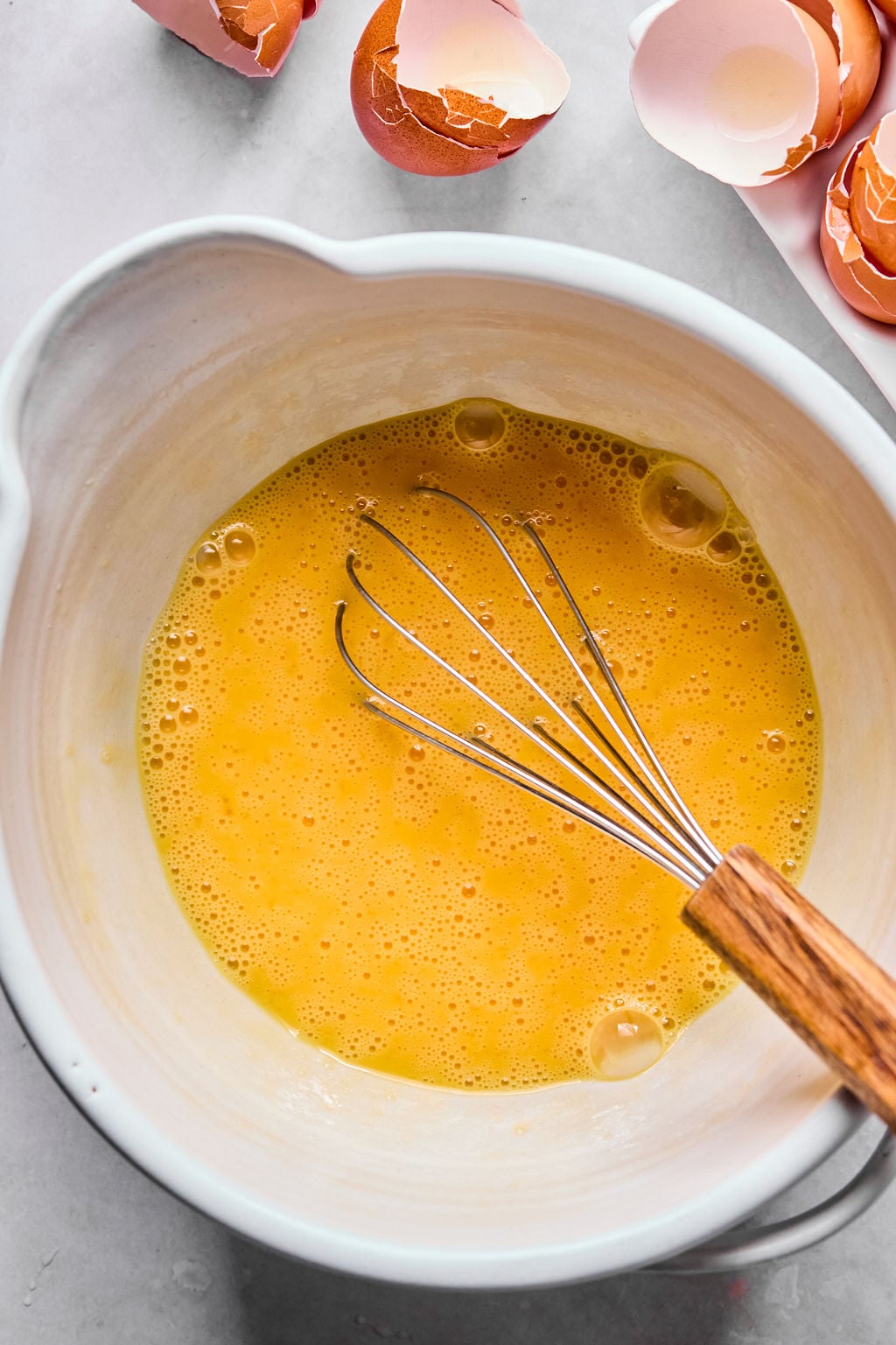 A whisk rests in a bowl of beaten eggs, with several cracked eggshells on the surrounding countertop.