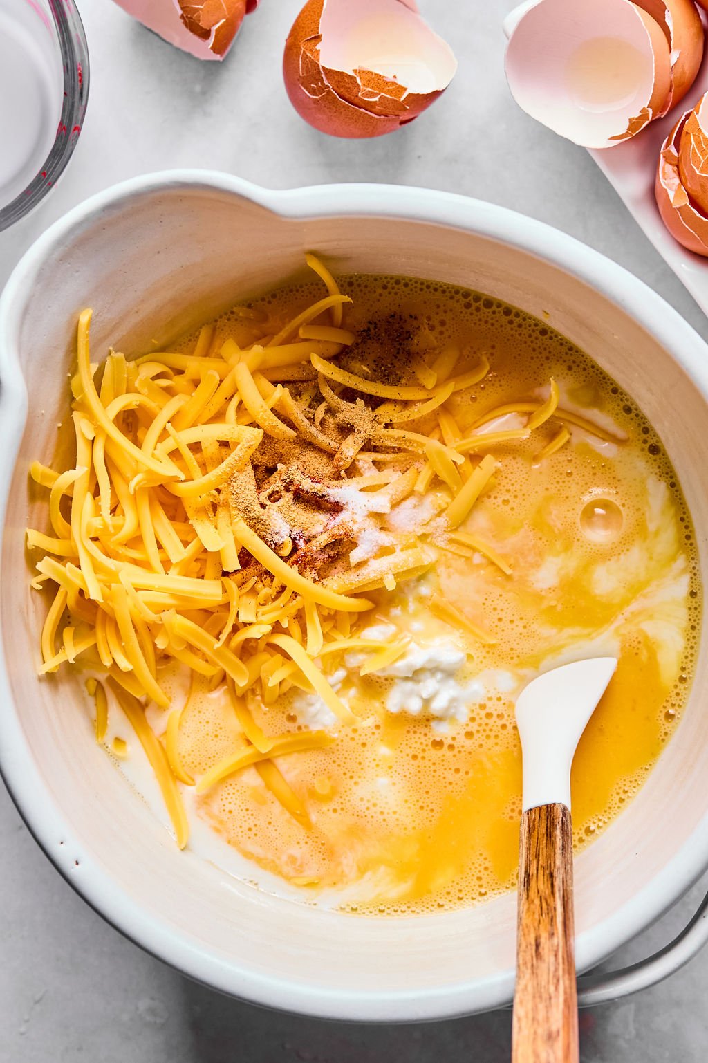 A mixing bowl with beaten eggs, shredded cheese, cottage cheese, spices, and a spatula, surrounded by broken eggshells.