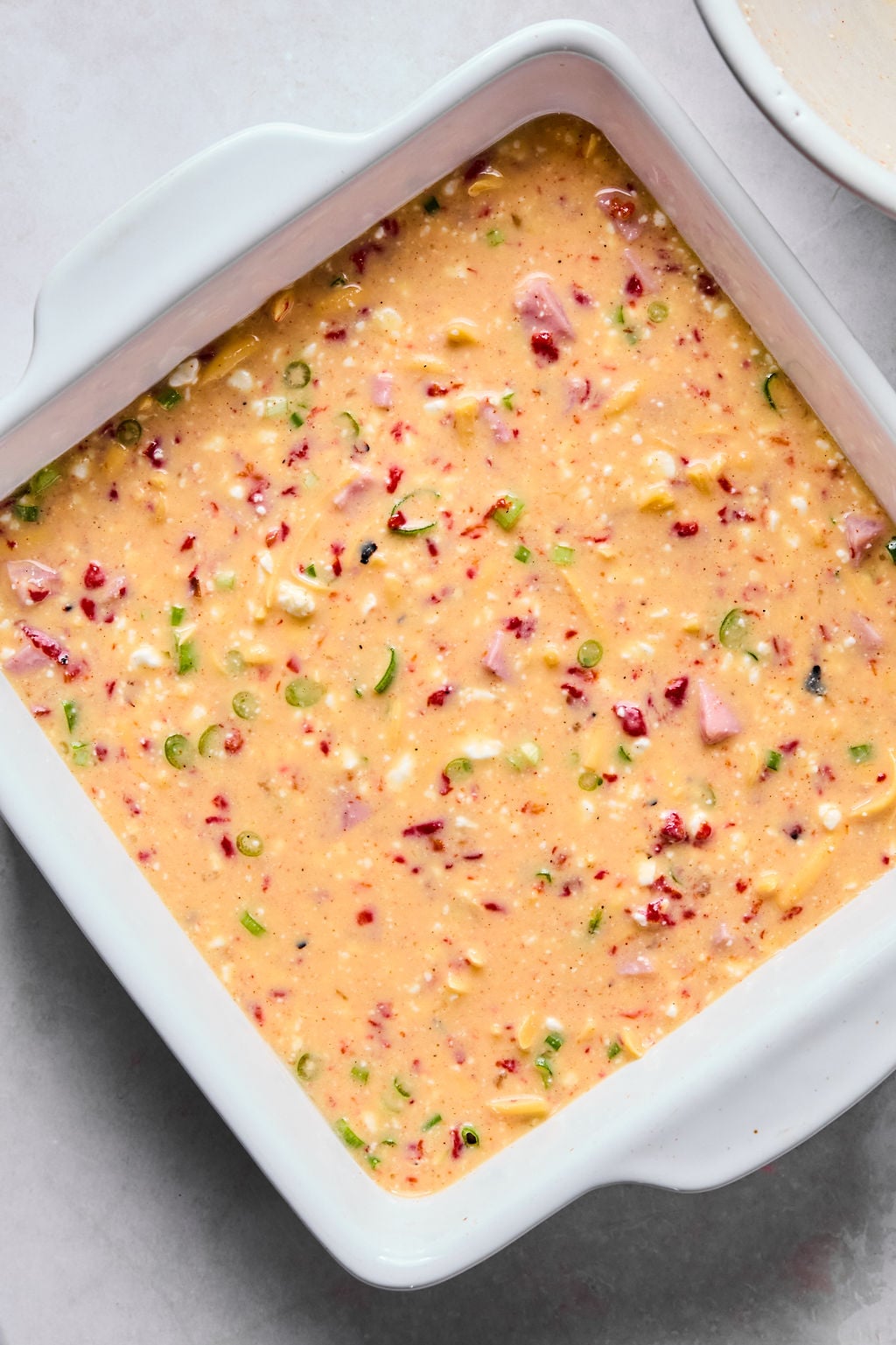 A square white baking dish filled with uncooked savory batter containing diced vegetables, ham, and cheese.