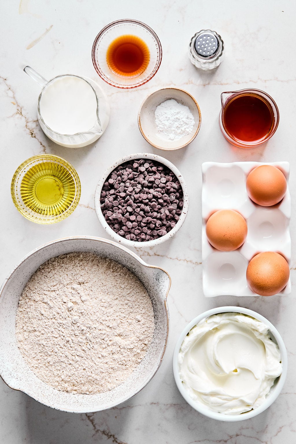 Assorted baking ingredients including flour, chocolate chips, eggs, vanilla, oil, yogurt, milk, syrup, baking powder, and salt arranged on a white countertop.