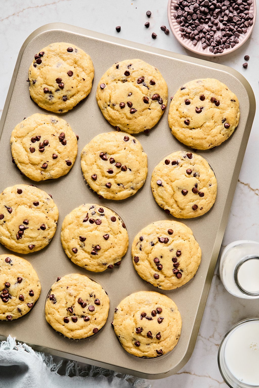A baking tray with twelve chocolate chip cookies, a small bowl of chocolate chips, and two glasses of milk on a marble surface.