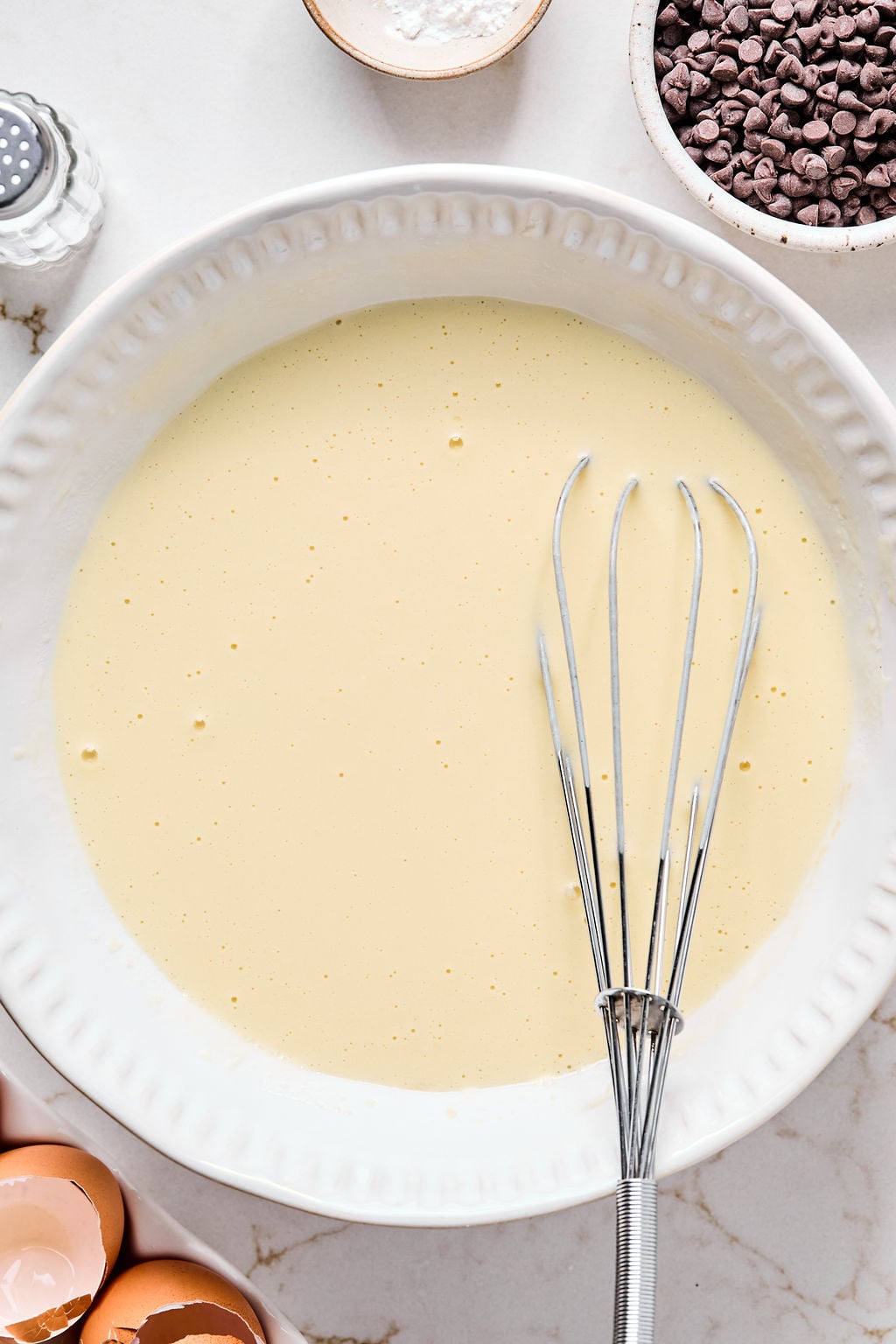 A white bowl filled with blended batter and a metal whisk, surrounded by chocolate chips, egg shells, and a small bowl of flour on a marble surface.