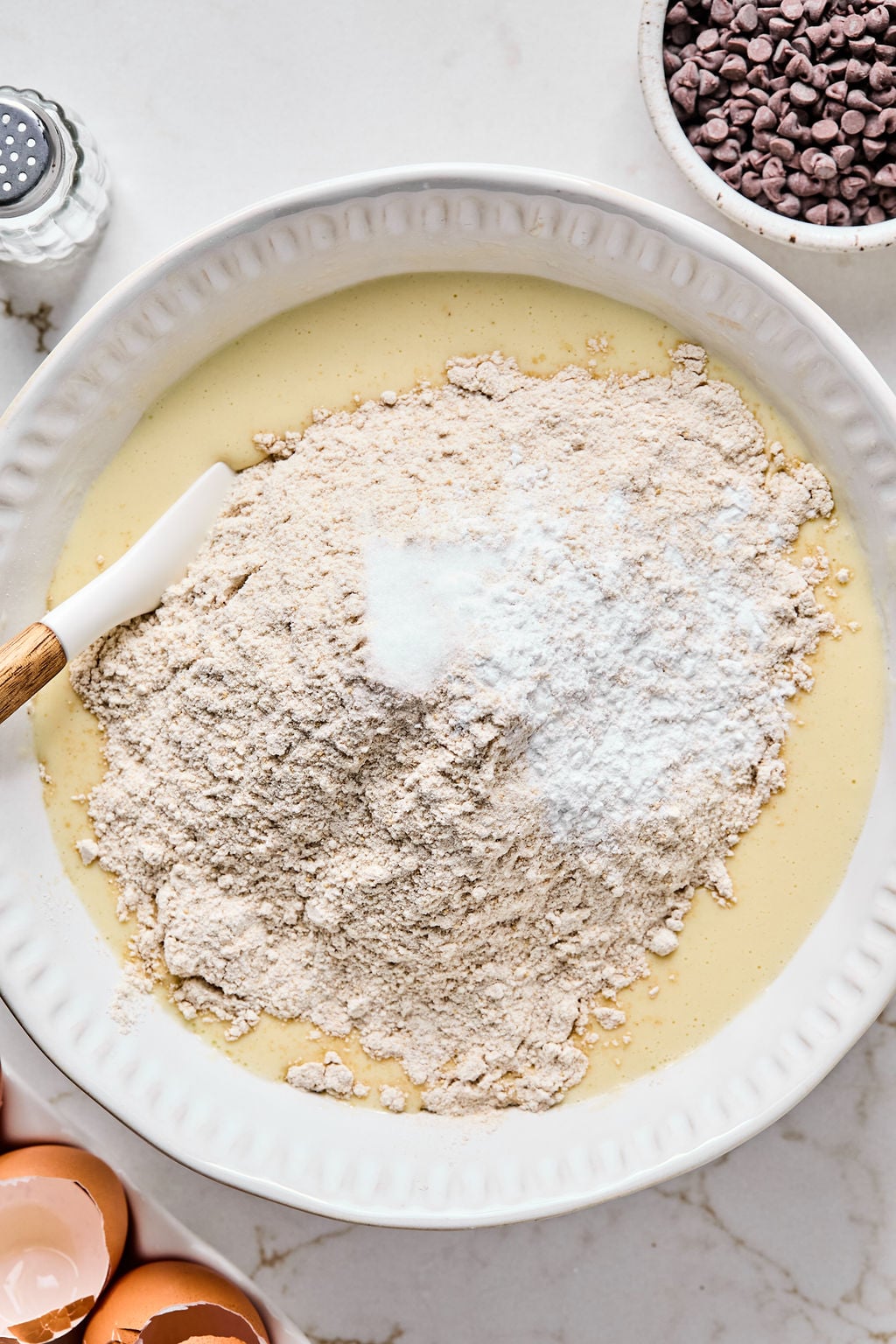 A white bowl with flour, sugar, and baking powder on top of a yellow batter, with a spatula in the mixture.