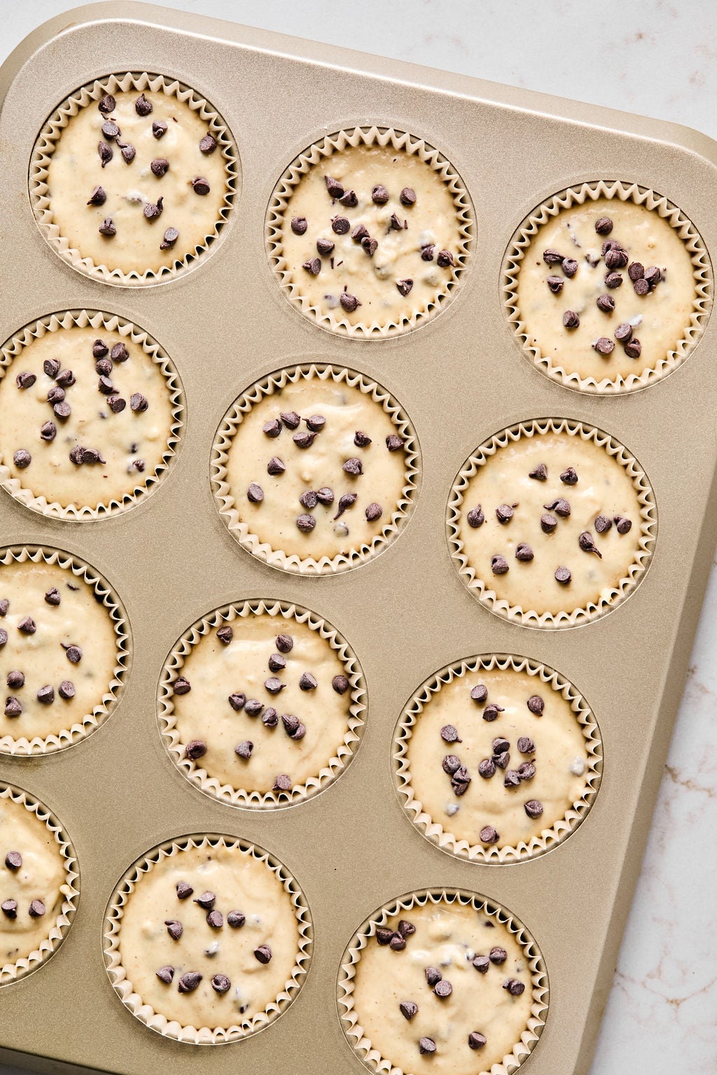 A muffin tin filled with unbaked muffin batter in paper liners, each topped with chocolate chips, sits on a light countertop.