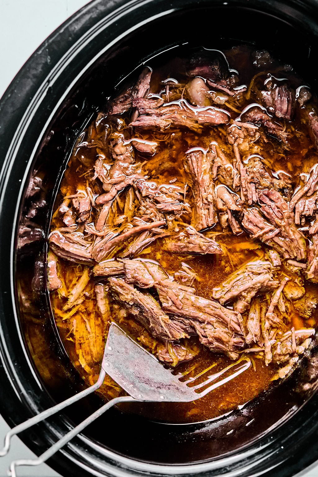Shredded beef in a slow cooker with broth, accompanied by a serving spatula and fork.