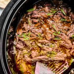 Shredded beef in sauce garnished with sliced green onions and sesame seeds inside a slow cooker, with serving forks placed on the side.