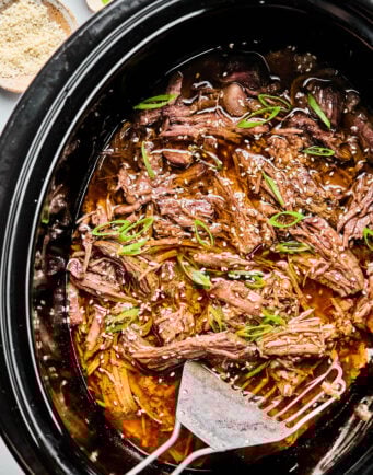 Shredded beef in sauce garnished with sliced green onions and sesame seeds inside a slow cooker, with serving forks placed on the side.