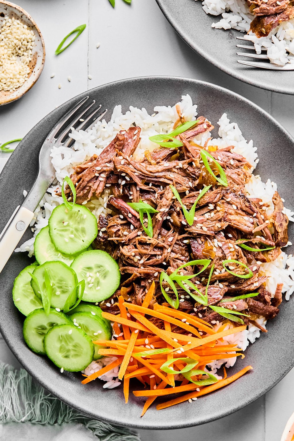 A gray plate with white rice, shredded beef, sliced cucumbers, julienned carrots, and green onions, garnished with sesame seeds, with a fork on the side.
