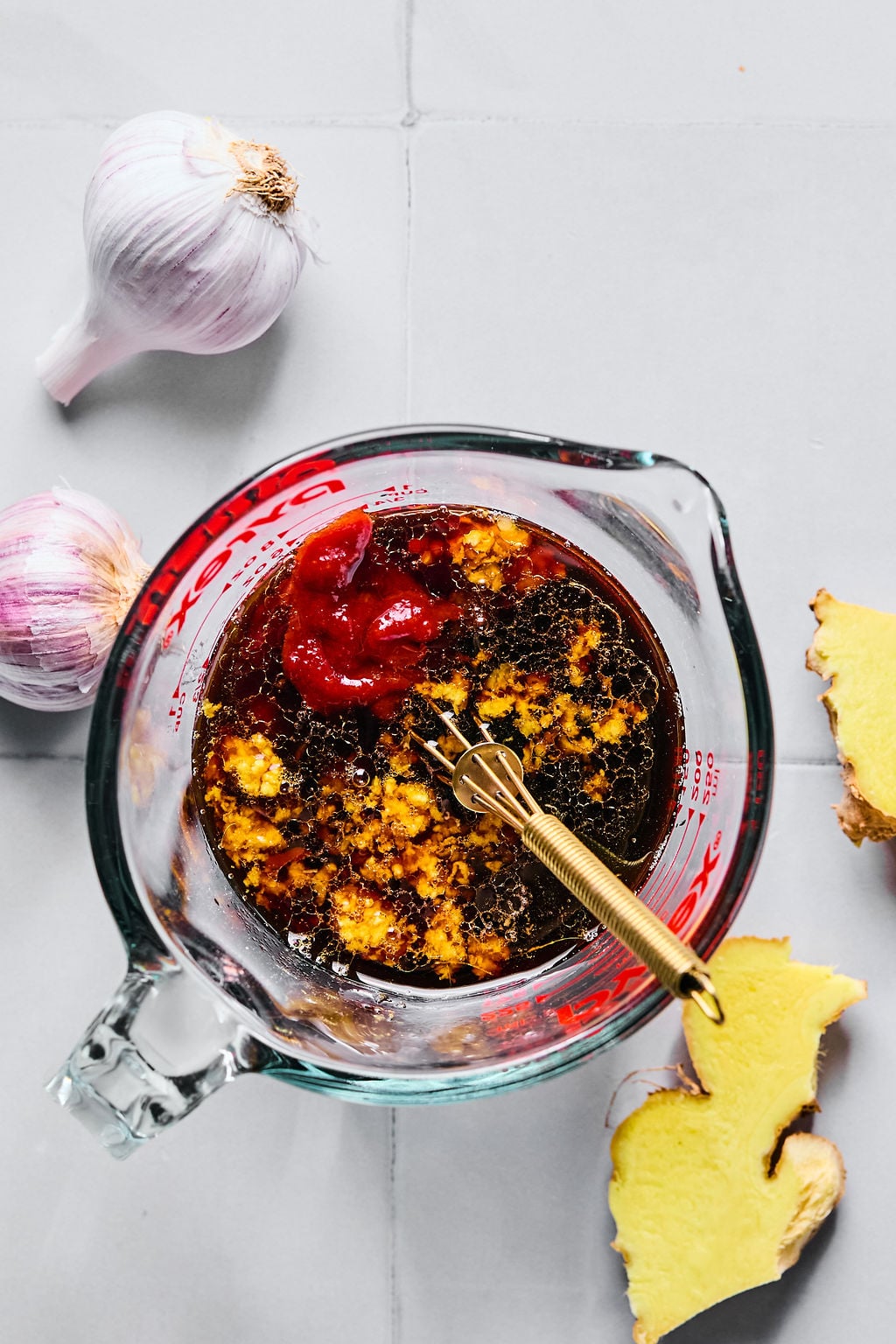 A glass measuring cup with a sauce mixture and gold whisk, surrounded by garlic bulbs and fresh ginger on a light gray surface.