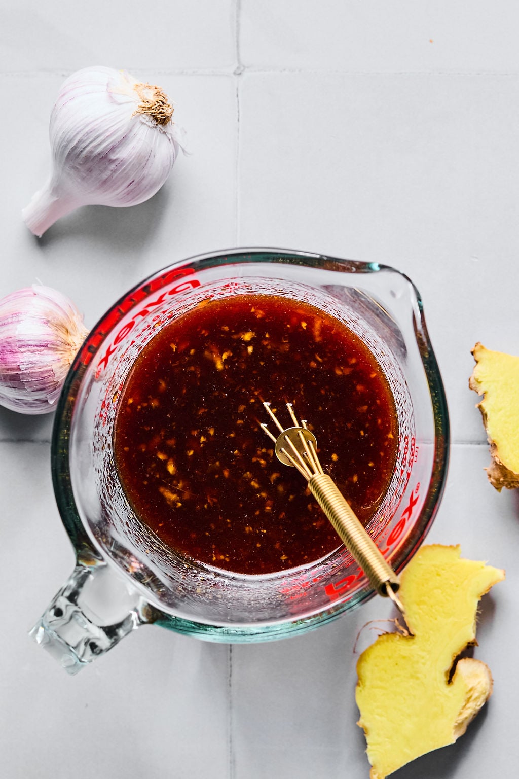 A glass measuring cup filled with a dark sauce and a small whisk, surrounded by garlic bulbs and slices of ginger on a light surface.
