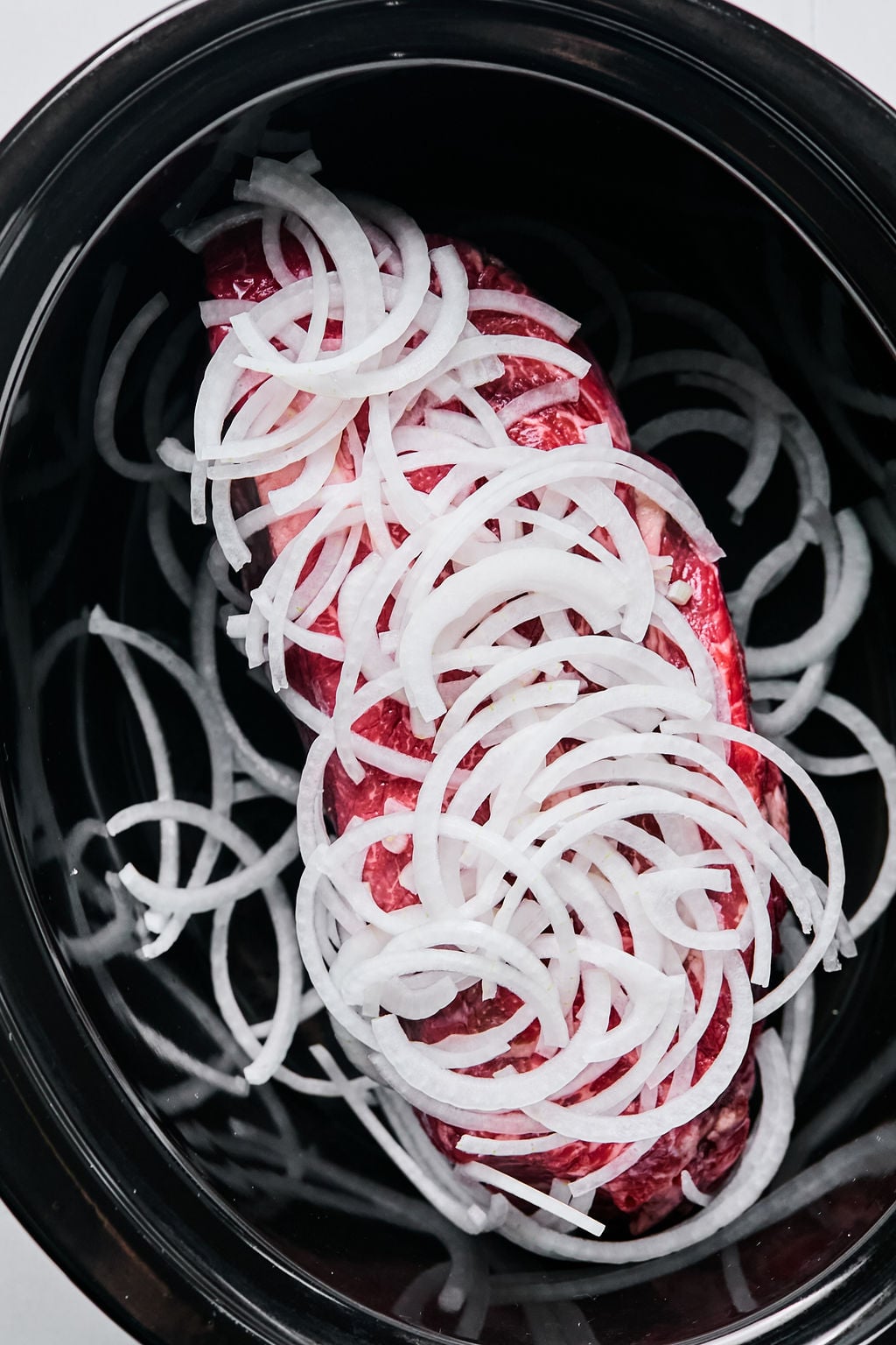 A raw piece of meat topped with sliced white onions in a black slow cooker.