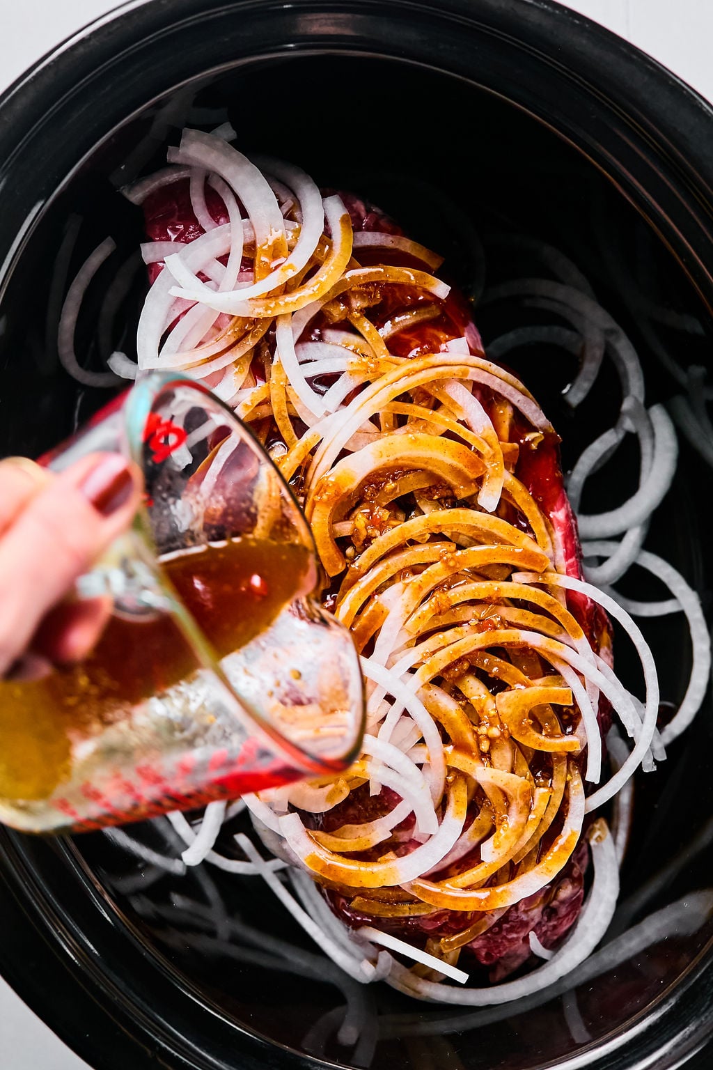 A hand pours a brown liquid over a piece of meat topped with sliced onions in a slow cooker.