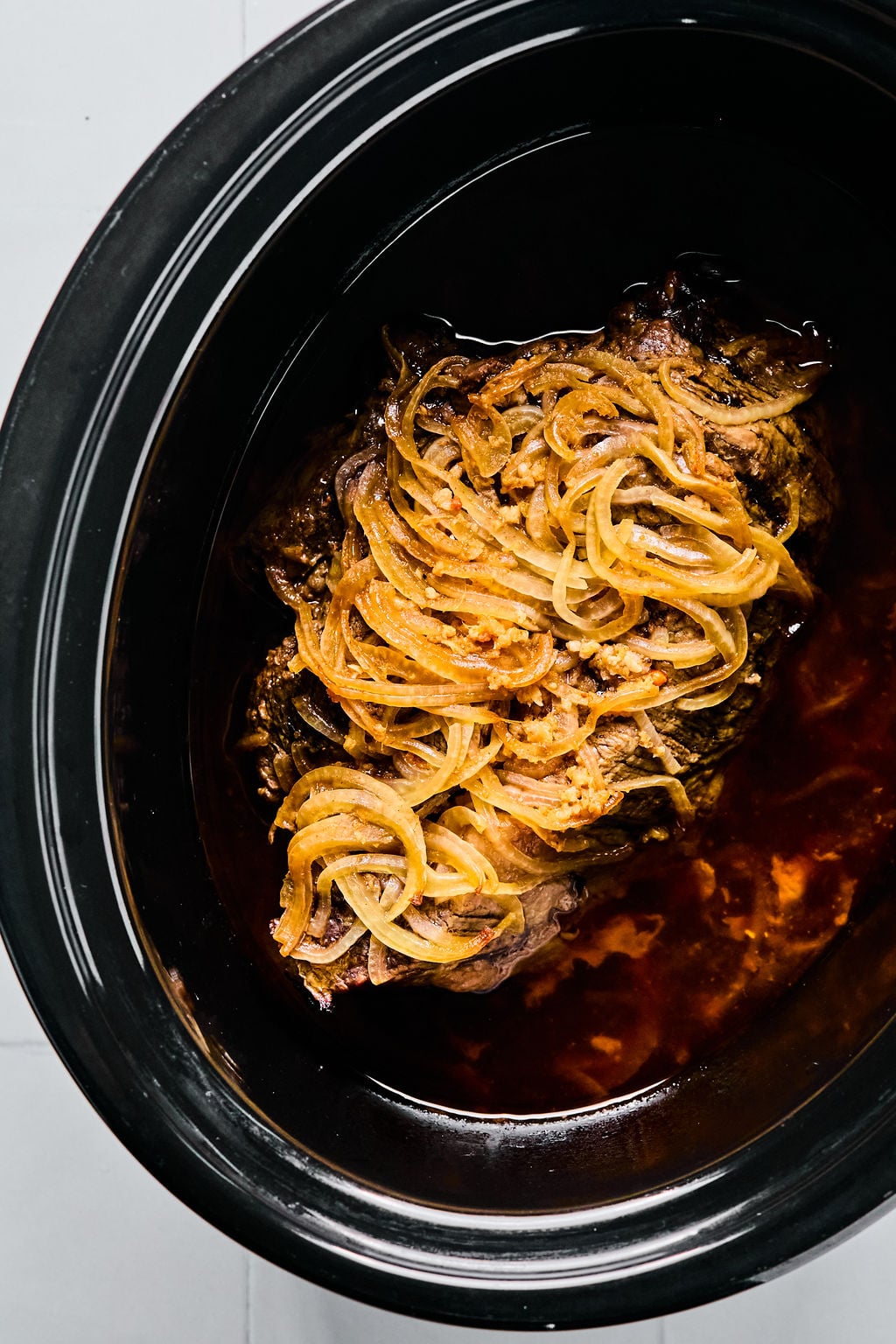 A cooked beef roast topped with sliced onions sits in a slow cooker with visible juices and sauce.